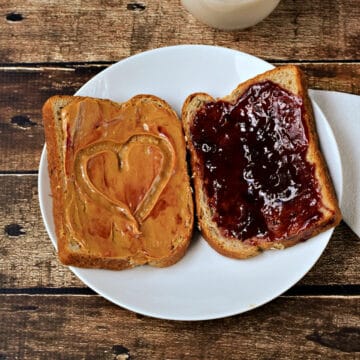Peanut butter on a slice of bread with a heart in the center and jelly on a second piece of bread sitting on a plate.