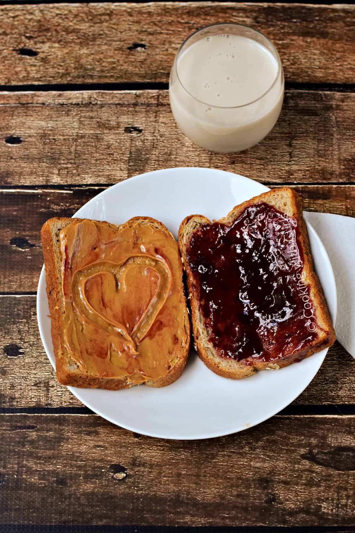 Looking down on a white plate with one slice of bread covered in peanut butter with a smear of jelly and a heart shape, and one slice of bread spread with jelly, with a glass of milk in the background.