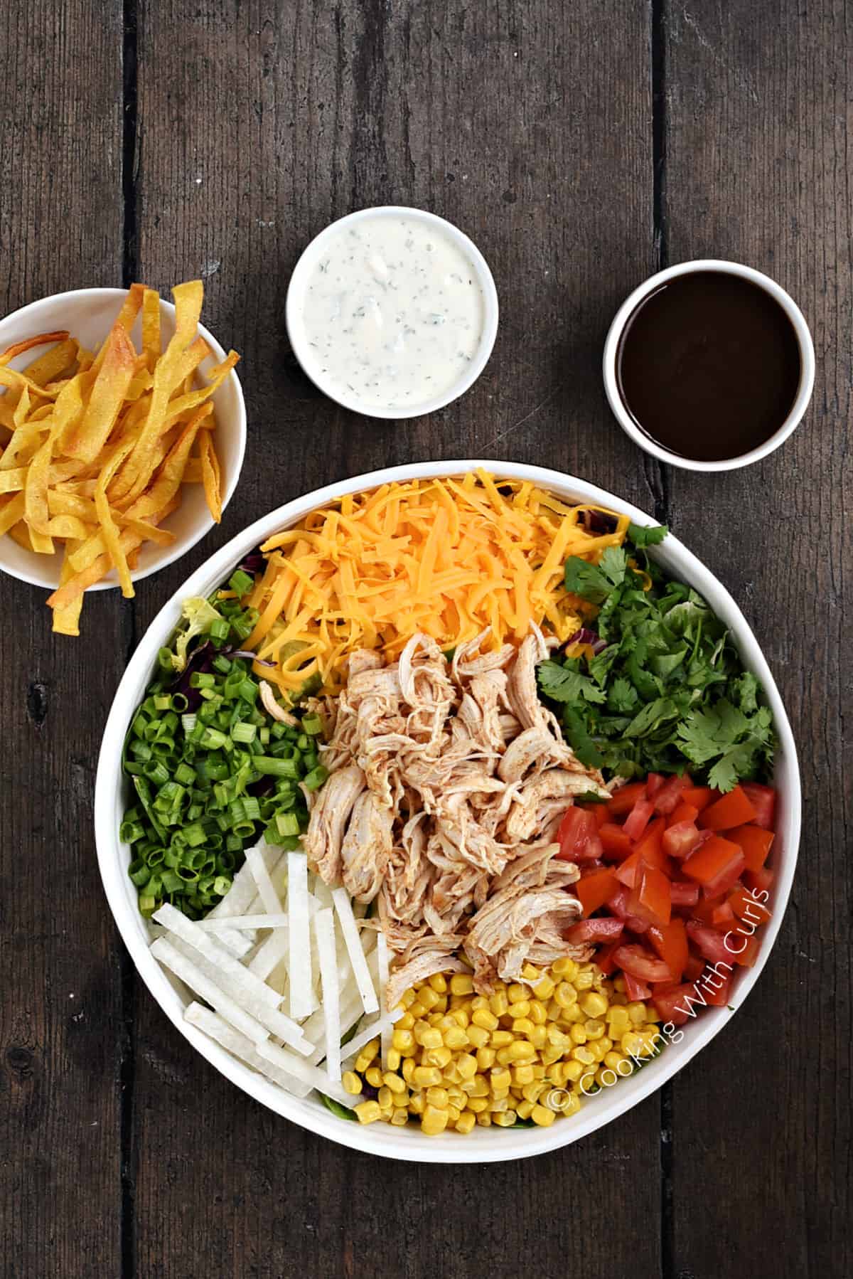 A large serving bowl filled with salad ingredients with bowls of tortilla strips, ranch dressing, and barbecue sauce in the background. 