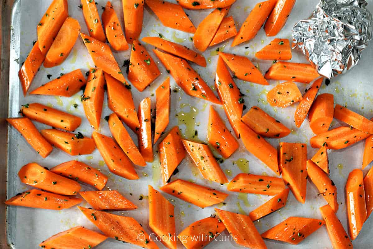 Chopped carrots tossed in oil and herbs on a baking sheet with foil wrapped garlic in the corner.
