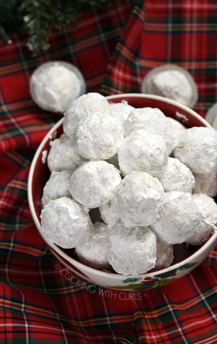 A bowl filled with Pecan Snowballs sitting on a red plaid napkin with two cookies in paper wrappers in the background.