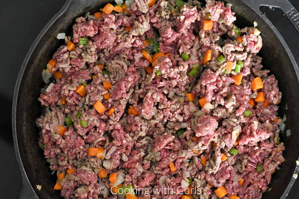 Raw ground beef mixed into diced vegetables in a cast iron skillet. 