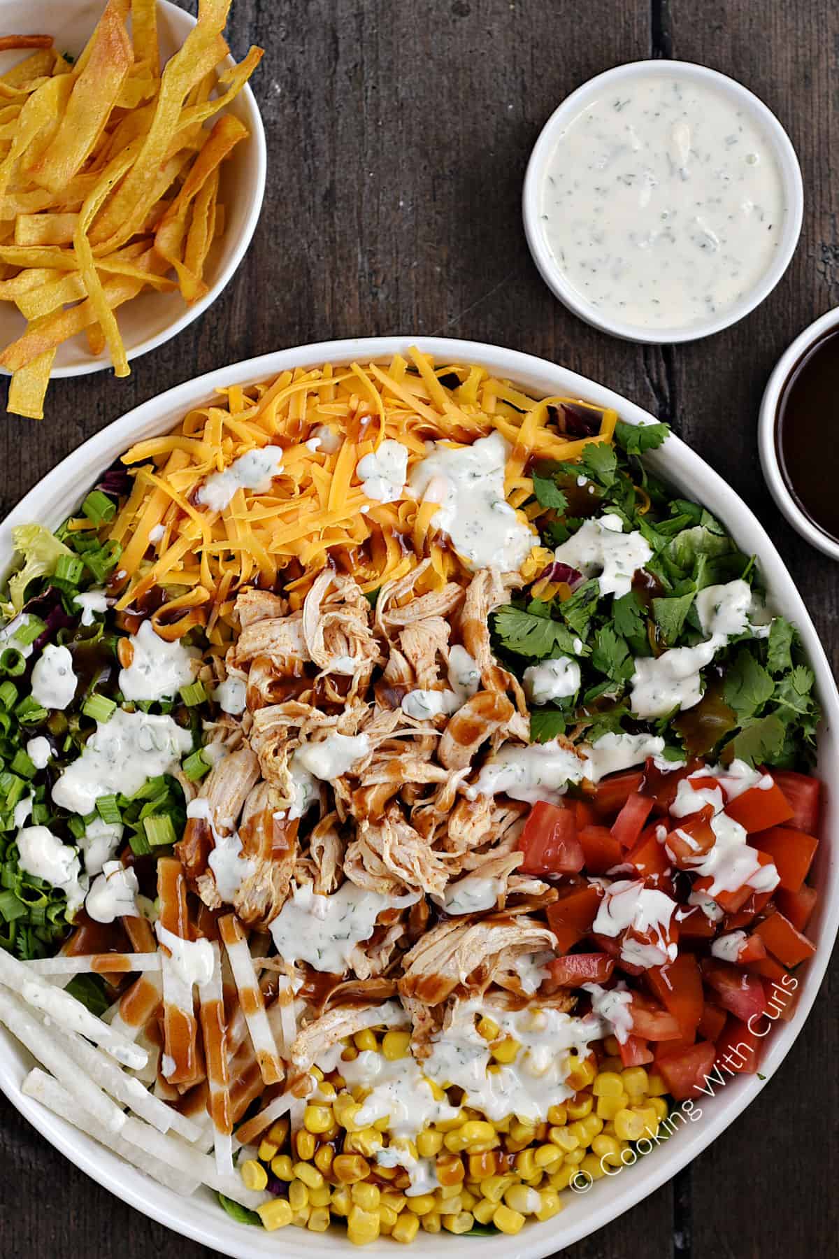 Salad ingredients in a large serving bowl drizzled with barbecue sauce and ranch dressing with tortilla strips and extra sauce in the background. 