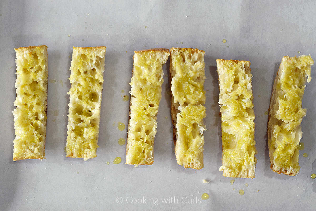 Six slices of ciabatta bread drizzled with oil on a baking sheet.