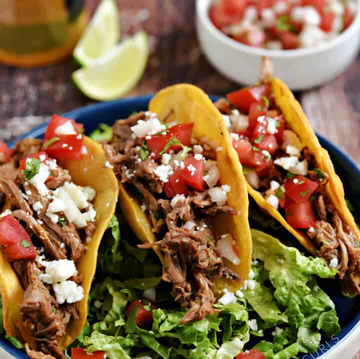 Three Mexican Shredded Beef hard-shell tacos on a bed of shredded lettuce with a bowl of pico, a bottle of beer and two lime wedges in the background.
