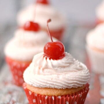three cupcakes with pale pink frosting topped with a single cherry lined up on a red, weathered wood board