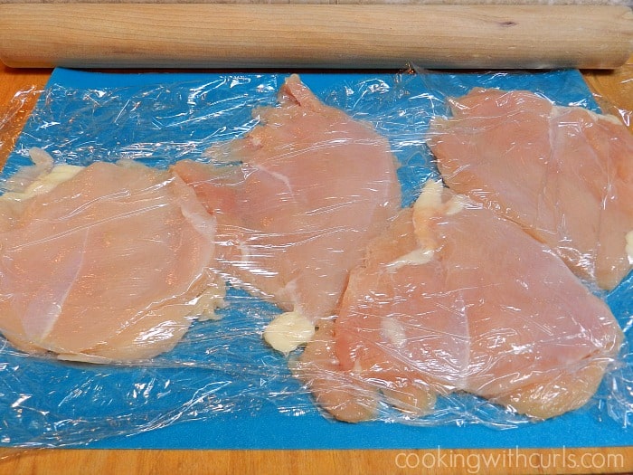 Four boneless, skinless chicken breasts on a wood board covered with plastic wrap and flattened with a rolling pin.