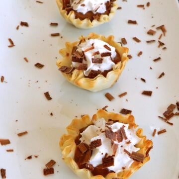 Four chocolate peanut butter pie bites lined up on a white platter sprinkled with shaved chocolate.