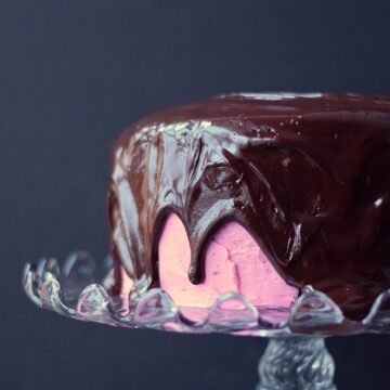 Raspberry Fudge Cake with chocolate ganache pouring over the top sitting on a glass cake stand.