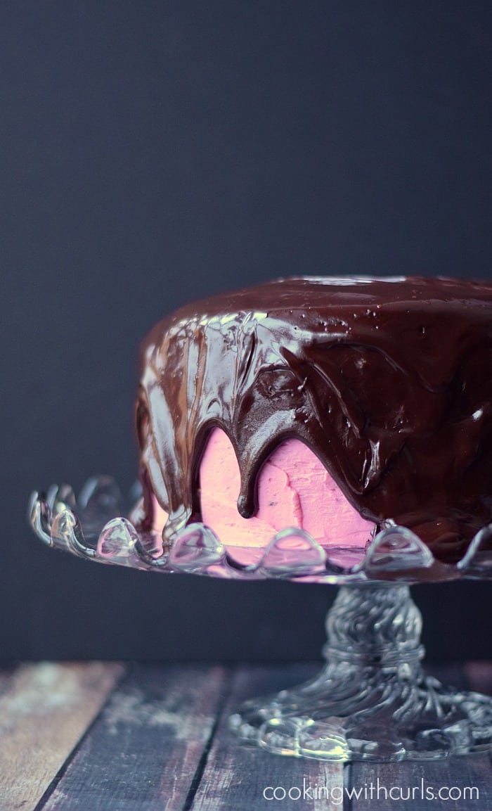 side image of a pink double layer cake with chocolate ganache dripping down the sides sitting on a crystal cake plate