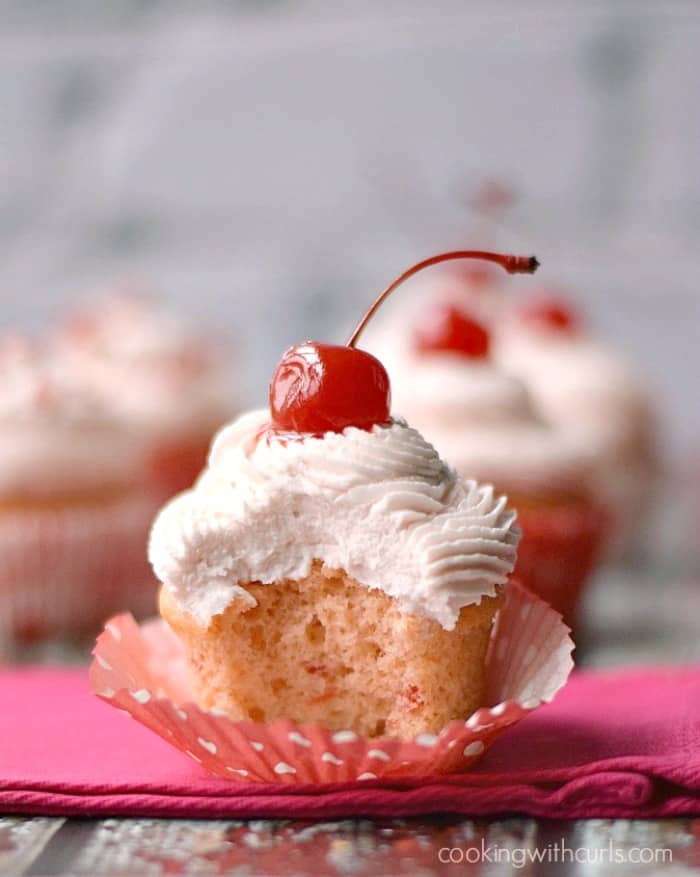 A cherry chip cupcake topped with pink frosting and a cherry with a big bite out of the front, sitting on a pink napkin with more cupcakes in the background.