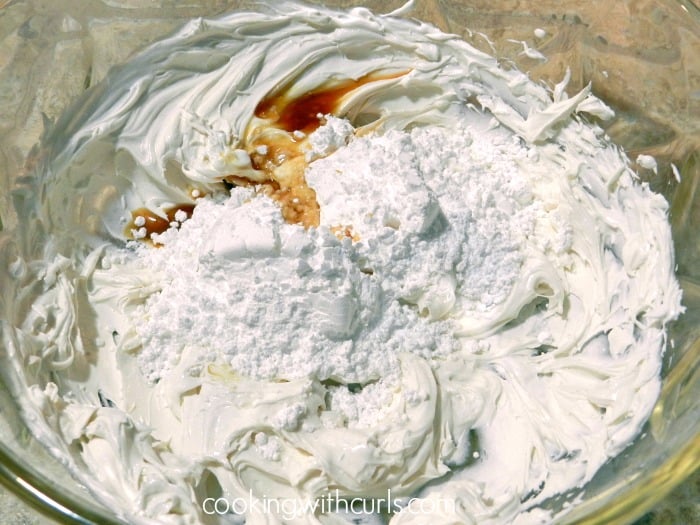 Vanilla and powdered sugar added to beaten cream cheese in a large glass bowl. 