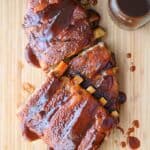 A slab of Guinness Barbecue Ribs on a wood cutting board with a jar of barbecue sauce in the upper right corner