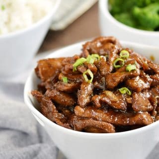 Mongolian Beef with thinly sliced sirloin, tangy Asian sauce, and sweet caramelized onions in a white bowl with rice and broccoli in the background
