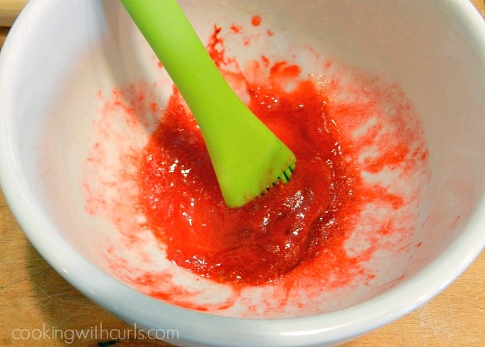 Red colored Egg Wash in a small white bowl mixed with a green silicone brush. 