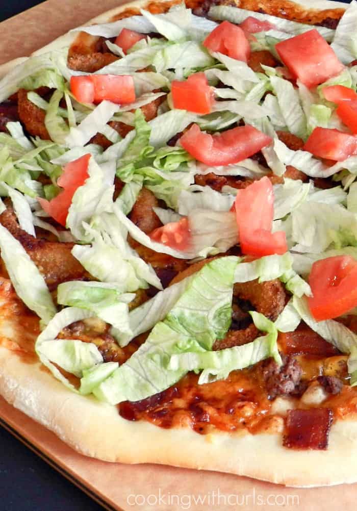 Bacon cheeseburger pizza topped with shredded lettuce and diced tomatoes sitting on a cutting board.