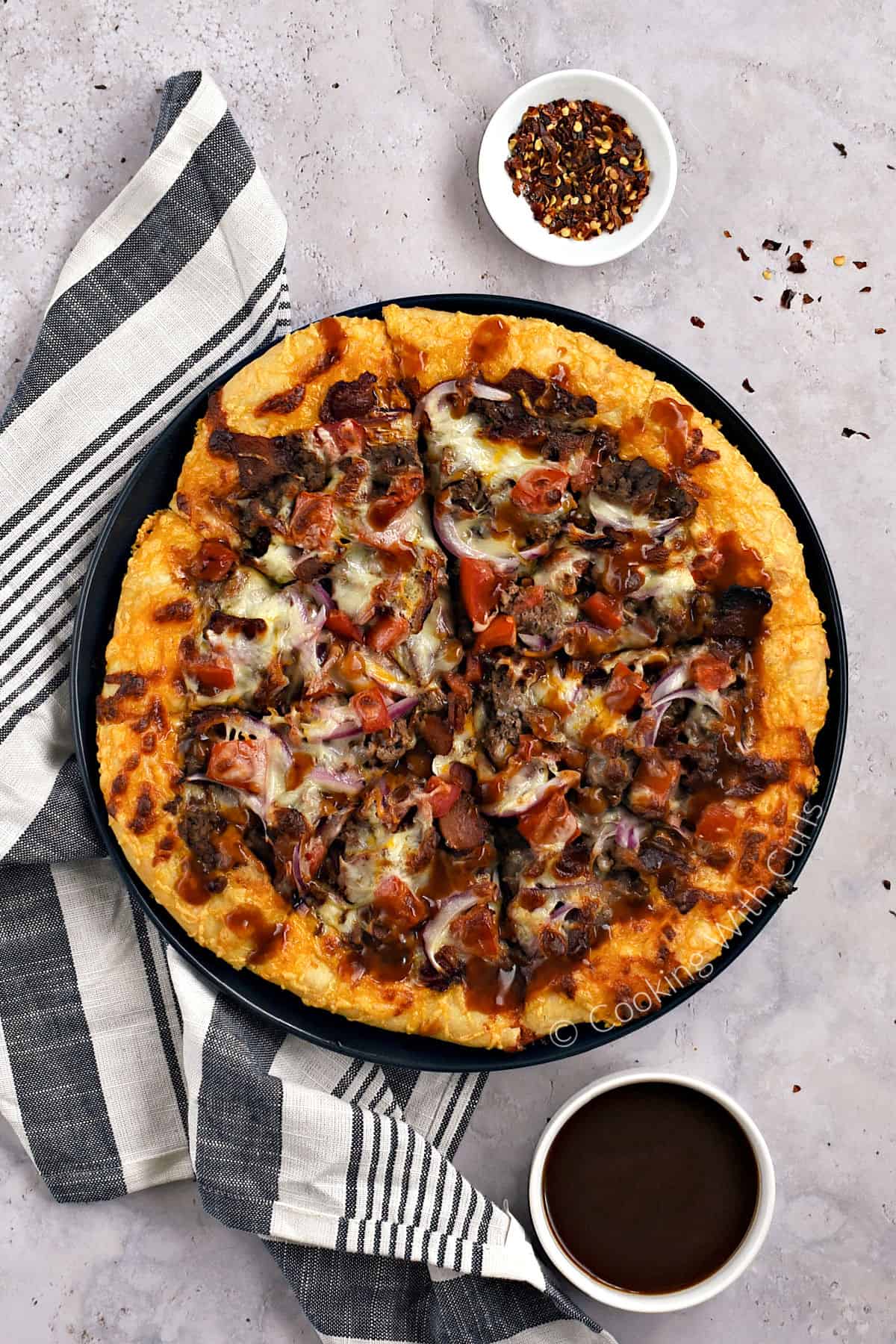 Looking down on a sliced bacon cheeseburger pizza cut into eight slices with chili flakes and barbecue sauce on the side.