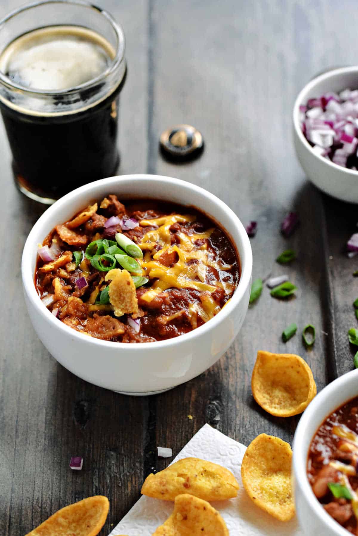 A bowl of beer chili topped with cheddar cheese, green onions, and sliced jalapeños with a glass of dark beer in the background.