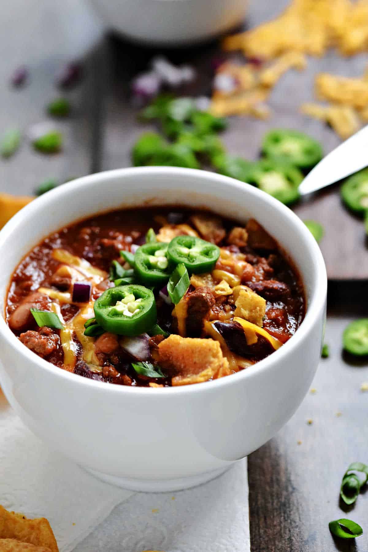 A bowl of beer chili topped with cheddar cheese, green onions, and sliced jalapeños.