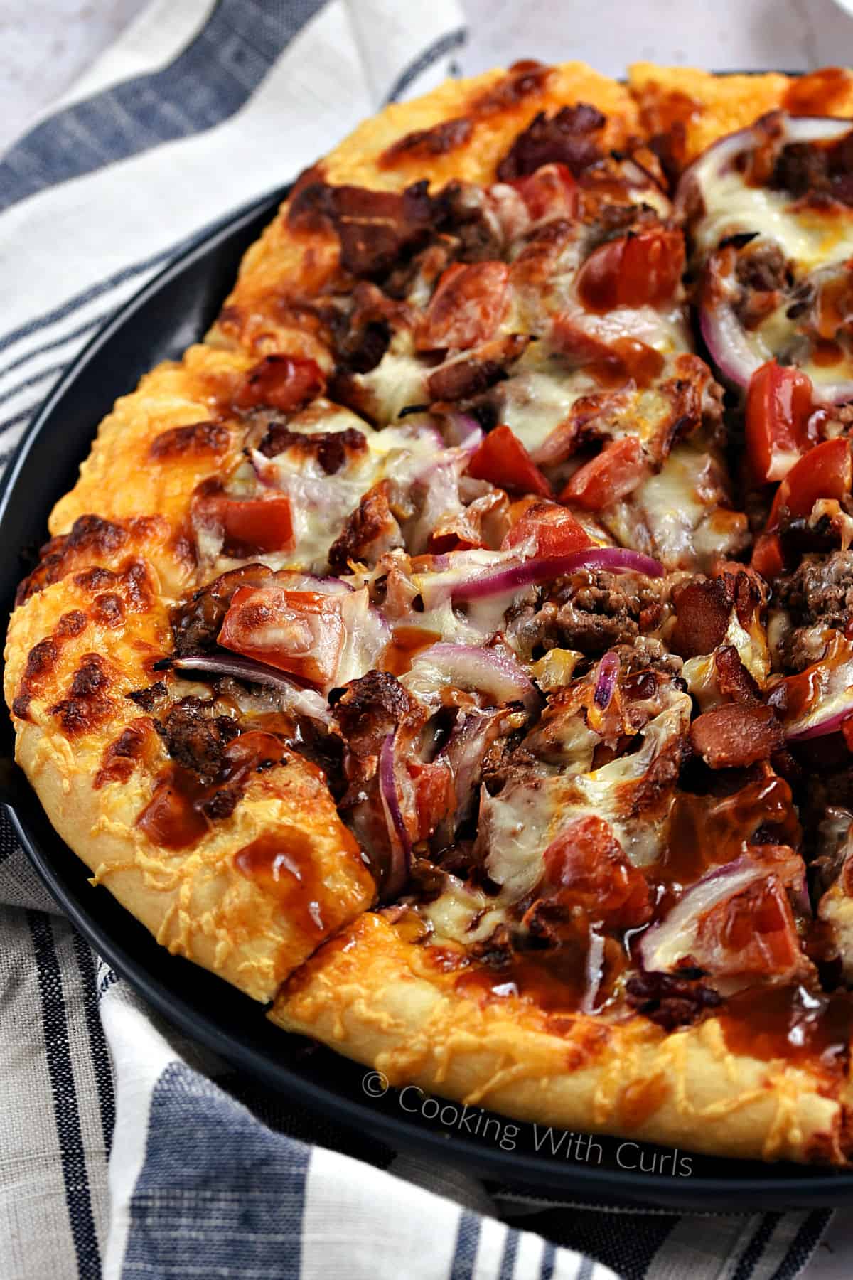 Close-up image of a sliced bacon cheeseburger pizza topped with cheese, beef, bacon, tomato, and barbecue sauce on a blue plate. 