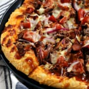 Close-up image of a sliced bacon cheeseburger pizza topped with cheese, beef, bacon, tomato, and barbecue sauce on a blue plate.