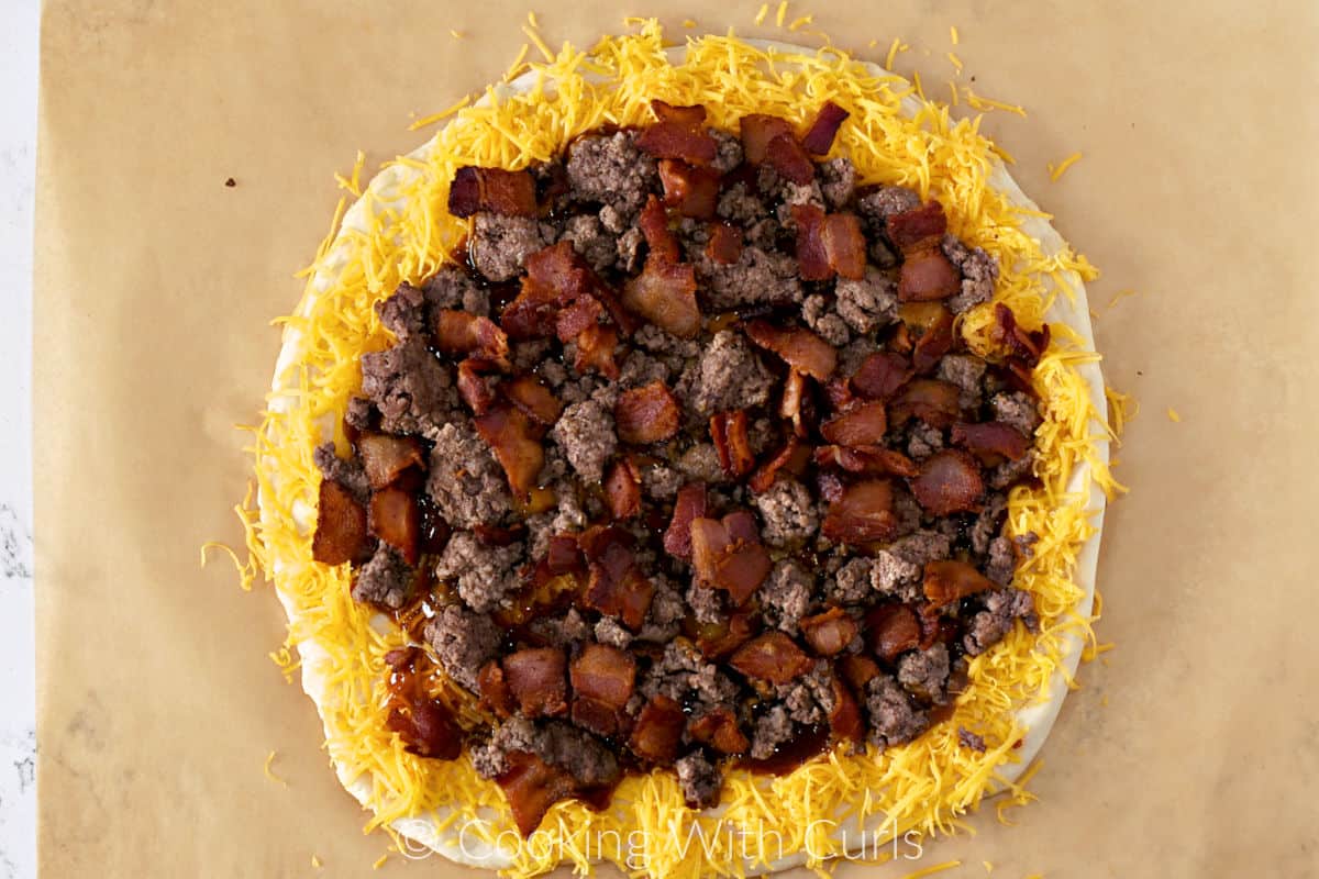 Cooked ground beef and bacon slices surrounded by grated cheddar cheese on a pizza dough circle. 