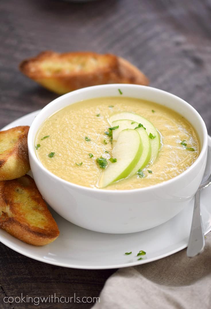 Creamy soup in a white bowl topped with green apple slices and green onions with crunchy bread slices on the side.