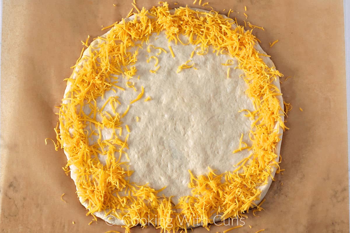 Pizza dough circle on a parchment paper with a ring of grated cheddar cheese around the edge. 
