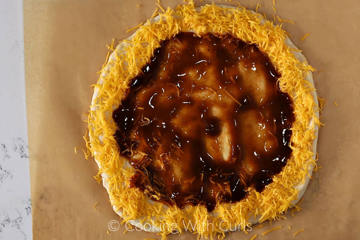 Pizza dough circle with barbecue sauce spread in the center and grated cheddar cheese around the edge. 