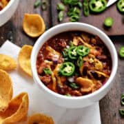 A bowl of beer chili topped with cheddar cheese, green onions, and sliced jalapeños.