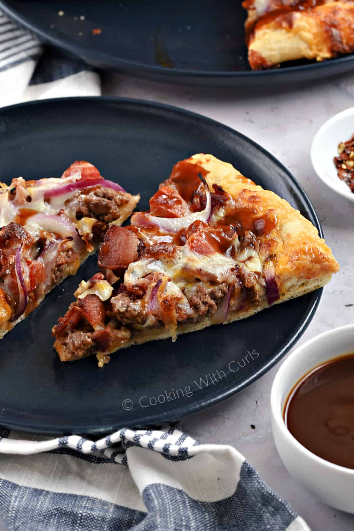 Two slices of bacon cheeseburger pizza on a blue plate with bowls of barbecue sauce and chili flakes on the side. 