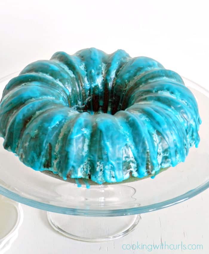 A Blue Kamikaze bundt cake on a glass cake stand with a white background 