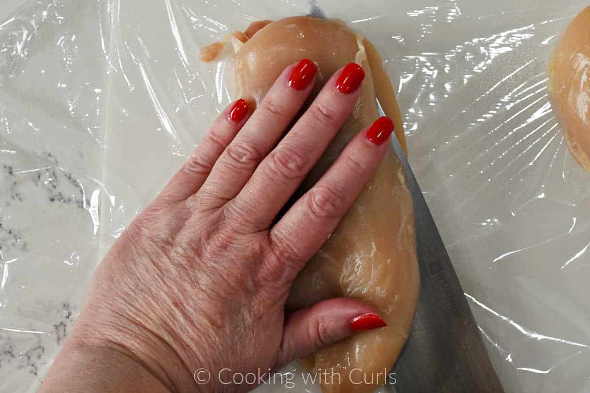 A woman's hand placed on a raw chicken breast while slicing it in half with a knife.
