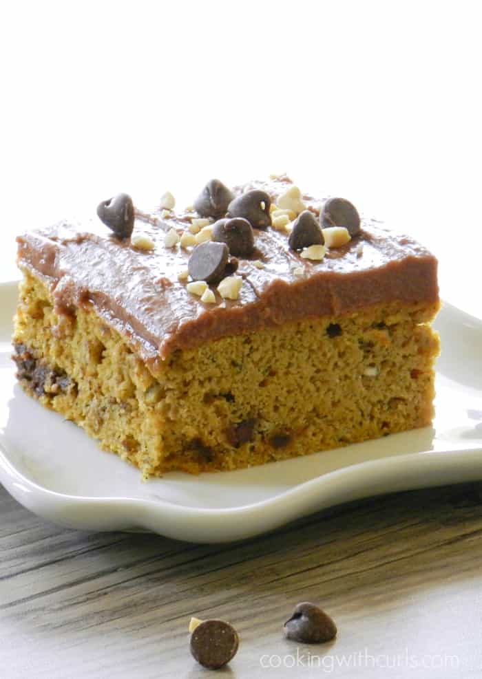 a slice of banana chocolate chip cake with chocolate peanut butter frosting topped with chocolate chips and chopped peanuts sitting on a square white plate