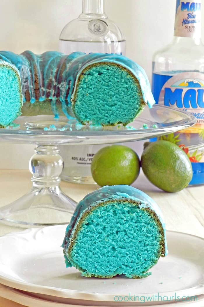 a slice of blue cake on a white plate with the remaining blue bundt cake on a glass cake stand with a bottle of vodka and bottle of blue Curacao in the background