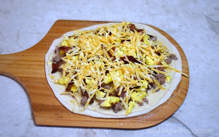 Grated cheddar cheese, scrambled eggs, bacon pieces, and sausage on top of gravy topped pizza dough.