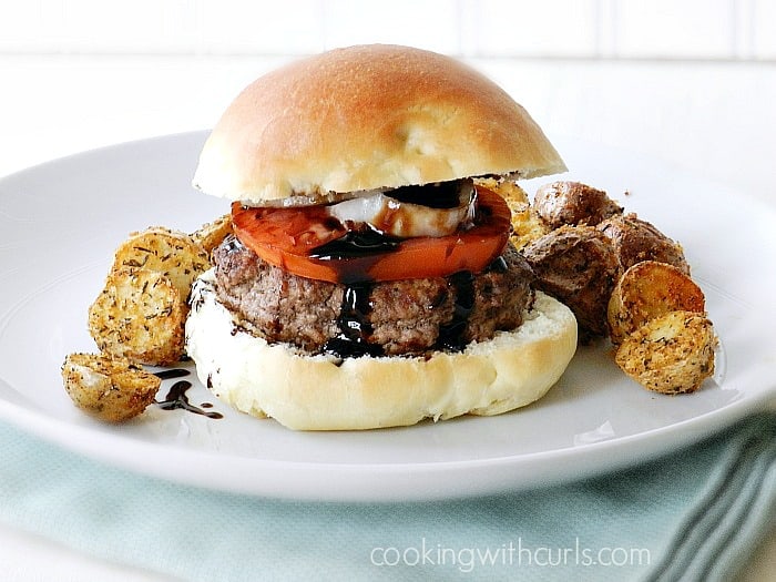 Greek Burger topped with onion, tomato and balsamic glaze surrounded by a bun sitting on a white plate with roasted potatoes