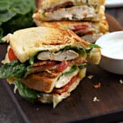 Two grilled chicken club sandwiches on golden sourdough bread with bacon, tomato, and ranch dressing stacked on a wood serving board.