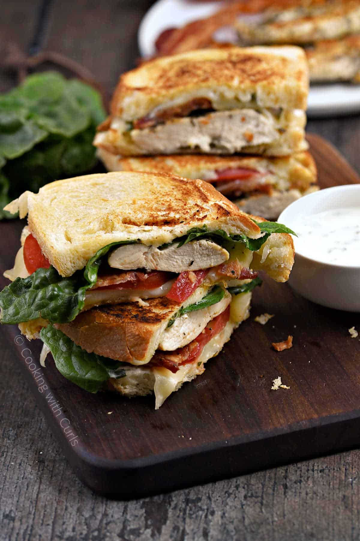 Juicy chicken breasts topped with ranch dressing, crispy bacon, tomato slices, and ranch dressing with grilled sourdough bread, cut in half and stack on a wood board with a second sandwich in the background.