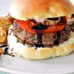 Ground lamb topped with balsamic glaze, onion and tomato surrounded by a hamburger bun sitting on a white plate with a light teal napkin underneath