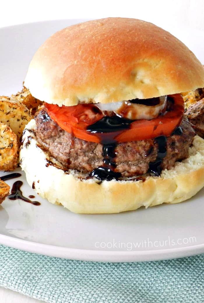 Ground lamb topped with balsamic glaze, onion and tomato surrounded by a hamburger bun sitting on a white plate with a light teal napkin underneath