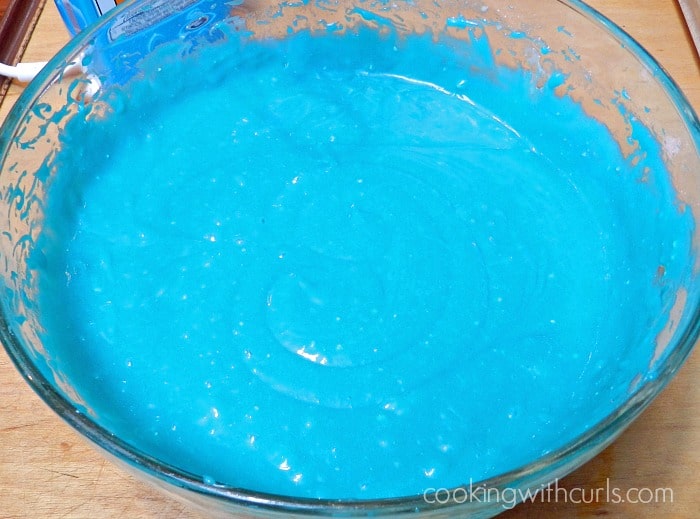 blue colored cake batter in a large glass bowl sitting on a wooden cutting board 