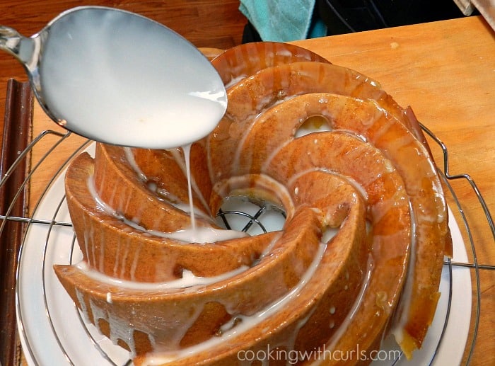 drizzle glaze over the baked cake sitting on a wire rack that is sitting on a large white plate