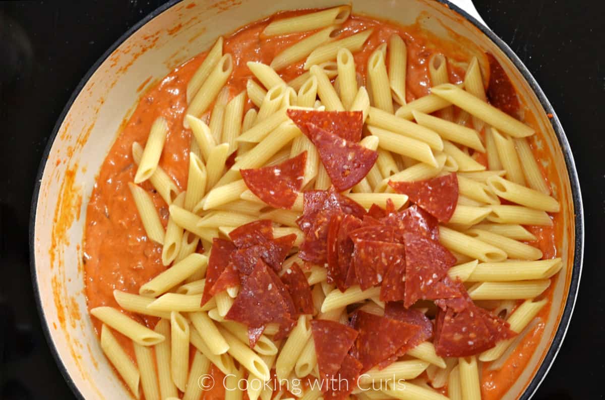 Cooked penne pasta, slices of pepperoni and creamy tomato sauce in a large skillet. 