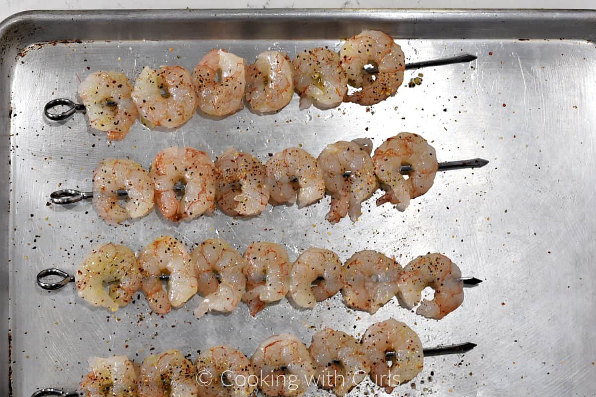 Four metal skewers each with 6 raw shrimp brushed with oil and seasoned with garlic pepper salt. 