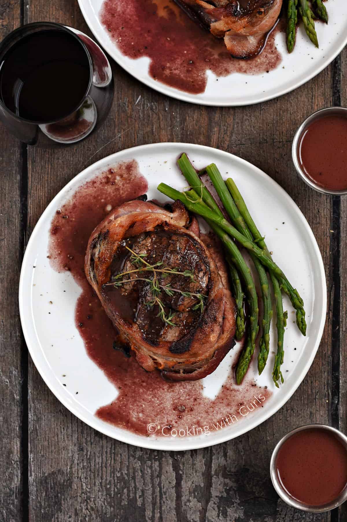 Looking down on bacon wrapped filet mignon with red wine sauce and asparagus spears on a white plate. 