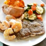 grilled steak, shrimp and vegetables on a large white plate with a baked sweet potato