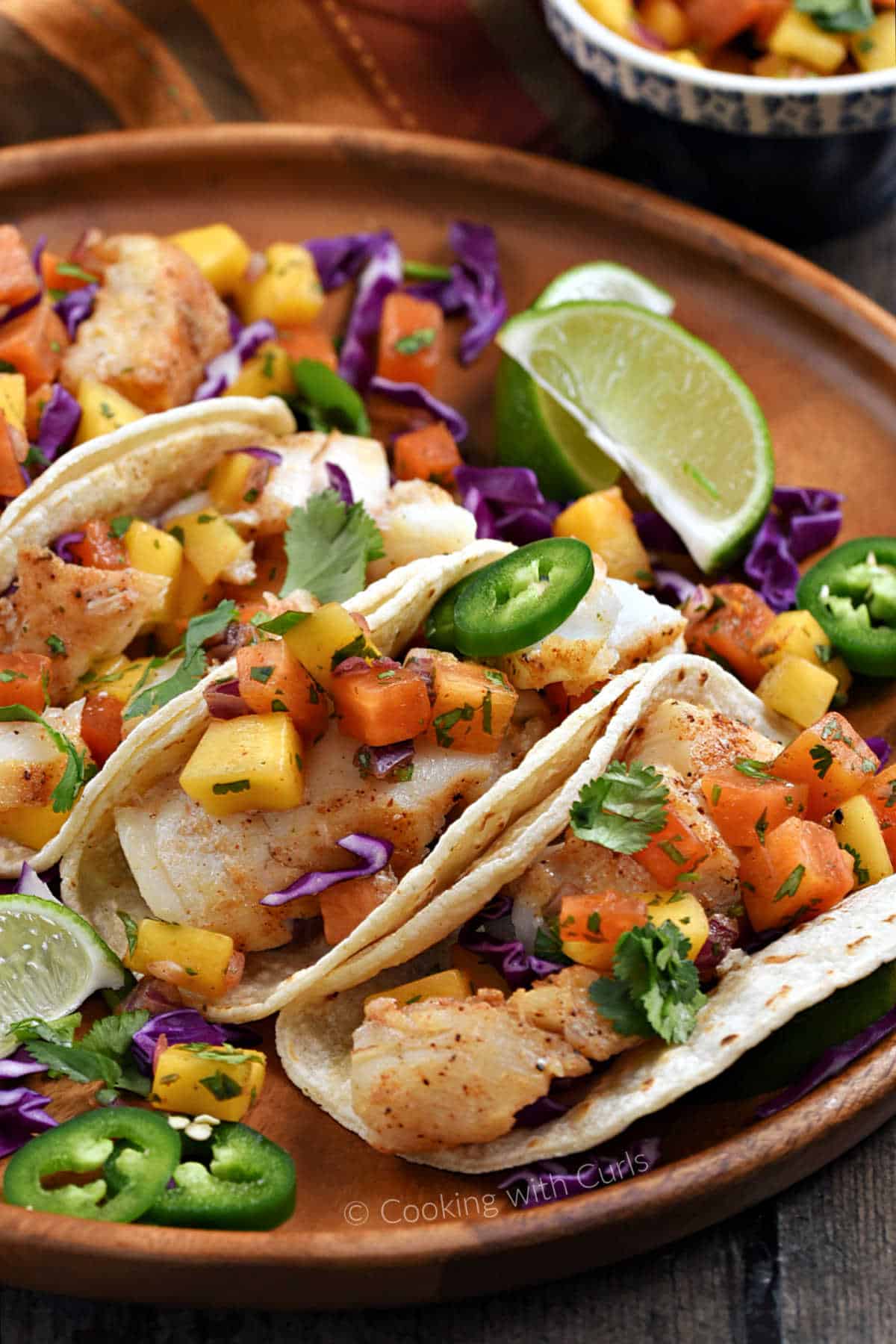 Three fish tacos with mango salsa and shredded red cabbage on a wood platter.