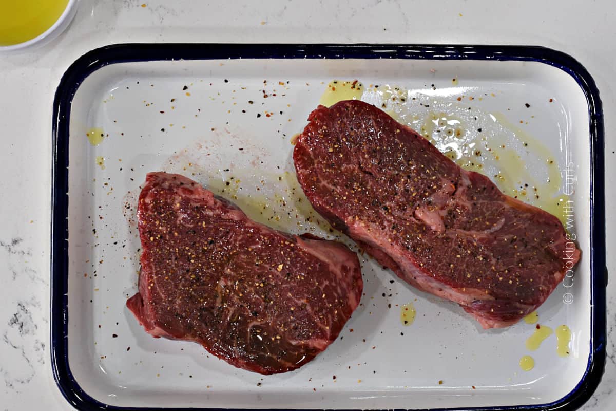 Two raw steaks on a platter topped with olive oil and garlic pepper seasoning salt. 