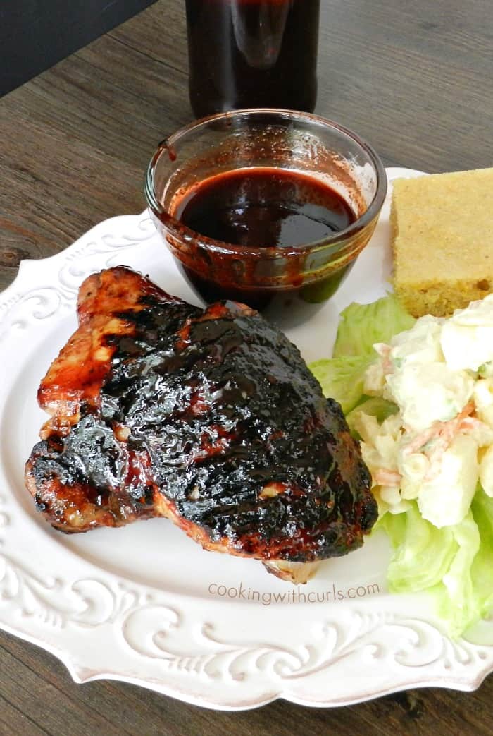 barbecue chicken, potato salad, cornbread and a bowl of barbecue sauce on a white plate with a bottle of sauce in the background